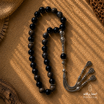 Natural black Obsidian stone Tasbih with polished beads and silver-tone metal details displayed on a textured brown background