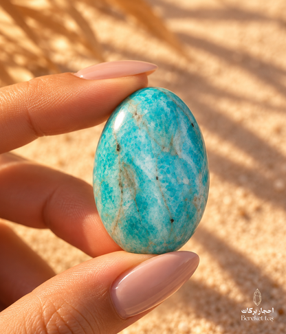 Natural polished Amazonite stone with blue-green tones held between fingers against a warm sandy background